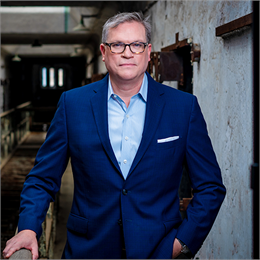 A headshot of Sean Kelly holding a handrail looking stoically at the camera wearing a suit and glasses