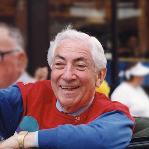 Ben Barkin rides in a vintage convertible waving happily to the crowd during a parade. His shirt is bright red and blue color block.